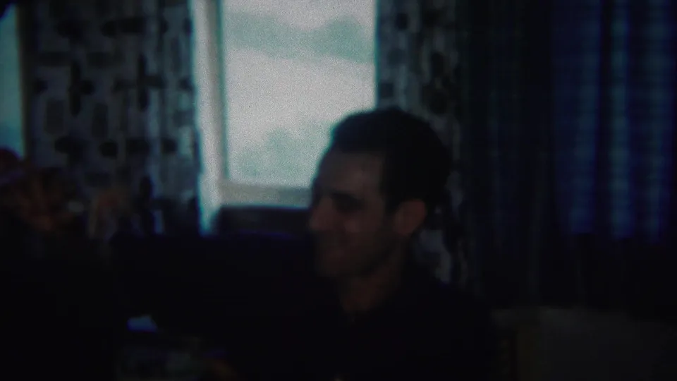 Adult man seated indoors during a 1951 Westville, New Jersey home movie meal, with patterned curtains and windows in the background.