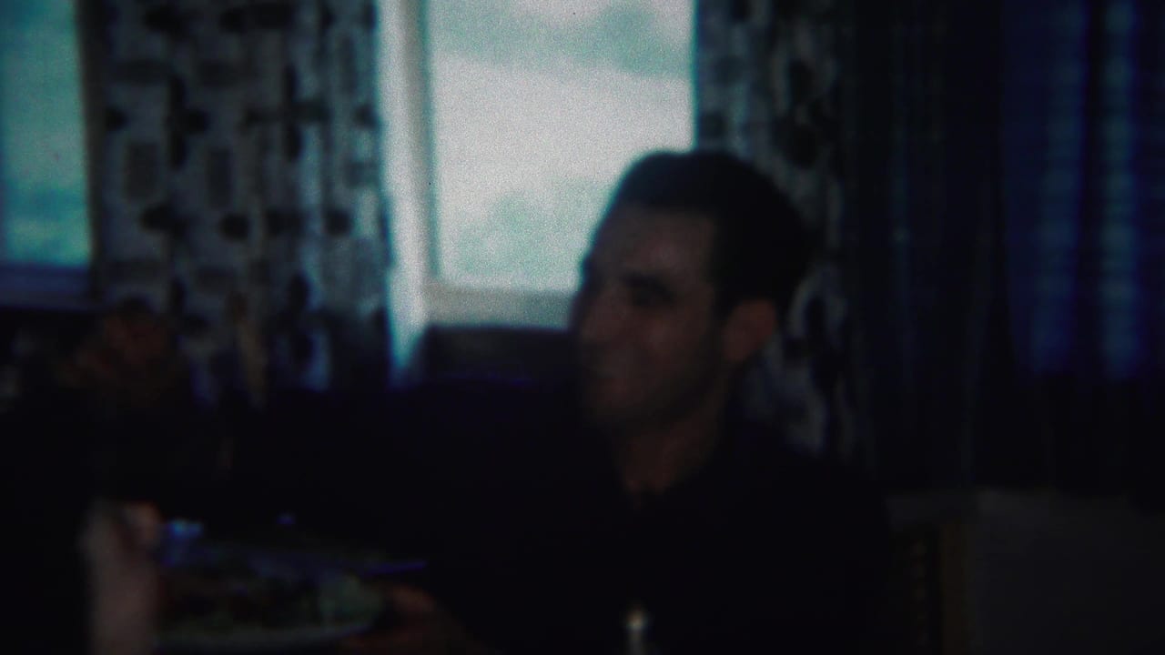 Man seated at a table in a dark interior with curtained windows behind him.