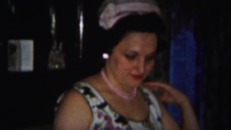 An adult woman in a patterned dress and pink headscarf appears seated indoors in the 1951 Westville home-movie dinner scene.