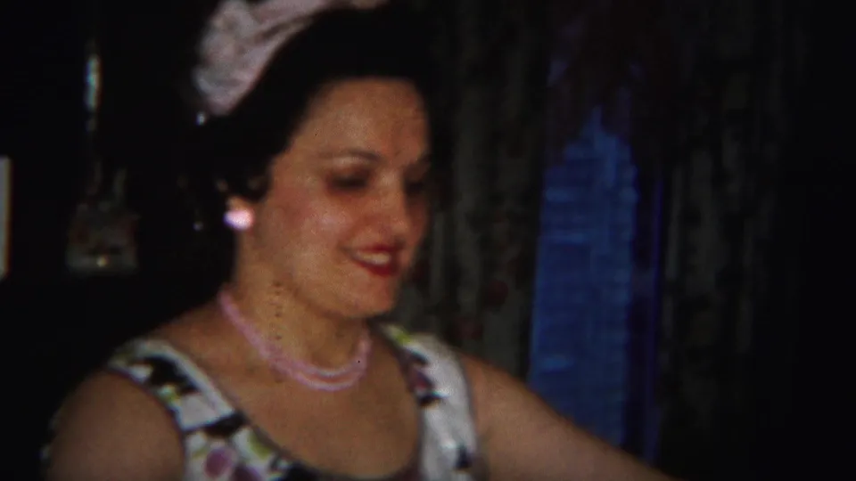 A smiling woman in a floral dress and pink necklace turns slightly right in the Westville, New Jersey 1951 home-movie interior.