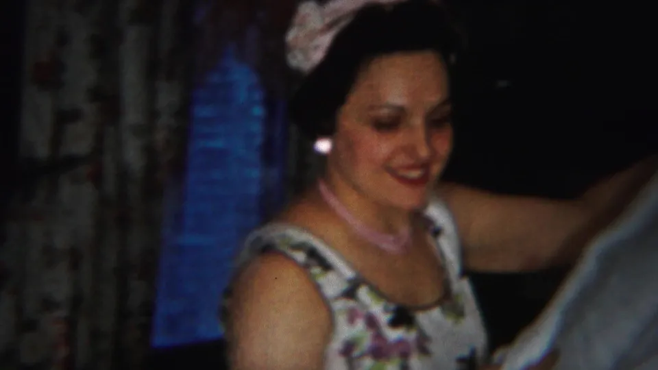 The woman smiles and lifts an arm near white lace fabric, with her floral dress, earrings, and pale headwear visible in 1951 Westville footage.