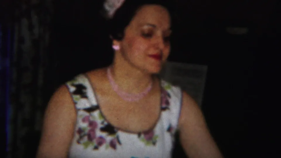 Adult woman in a floral dress and pink necklace in a dim Westville, New Jersey home-movie interior from the 1951 metadata.