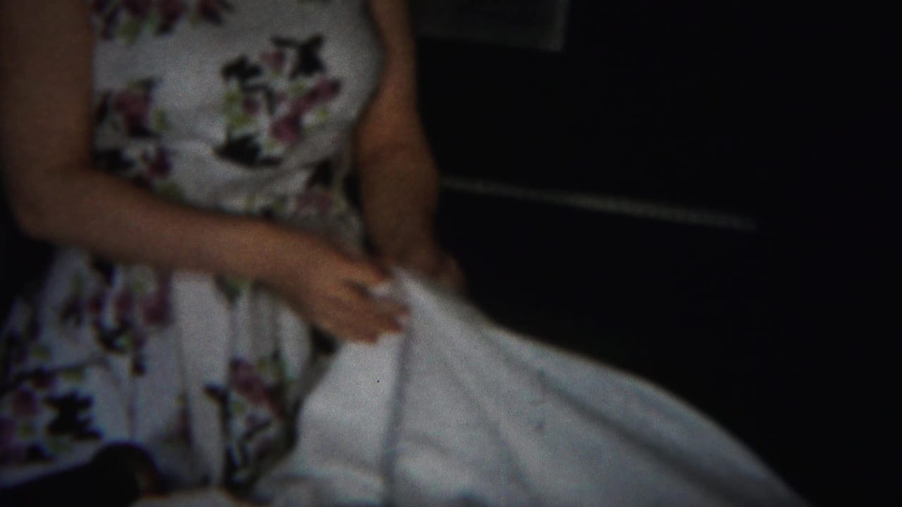 Woman in a floral dress holding a white sheet in a dark room.