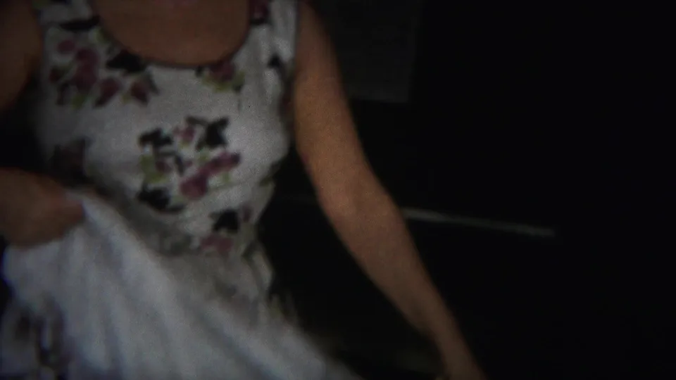 Close handheld view of the woman's floral dress and arm beside a white sheet in the domestic interior.