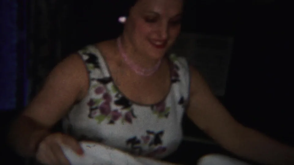 Smiling adult woman in a floral dress and pink necklace leans over a white sheet in a 1951 Westville home-movie interior.