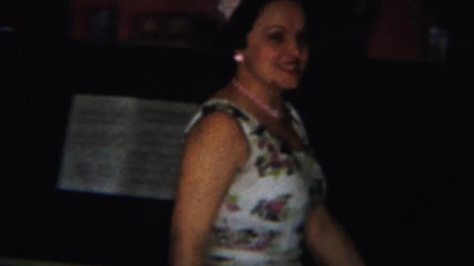 Adult woman in floral dress and jewelry is seen in a low-light home interior, with a possible music stand or piano area nearby.