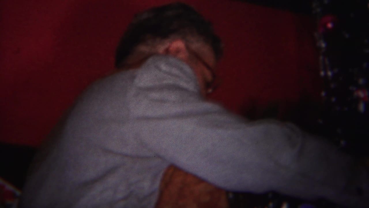 Man in a gray sweater reaches toward a tinsel-covered Christmas tree near a red wall.