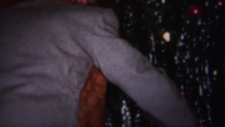 A close handheld frame shows the man's sweater and arm beside the Christmas tree, with tinsel catching small points of light.