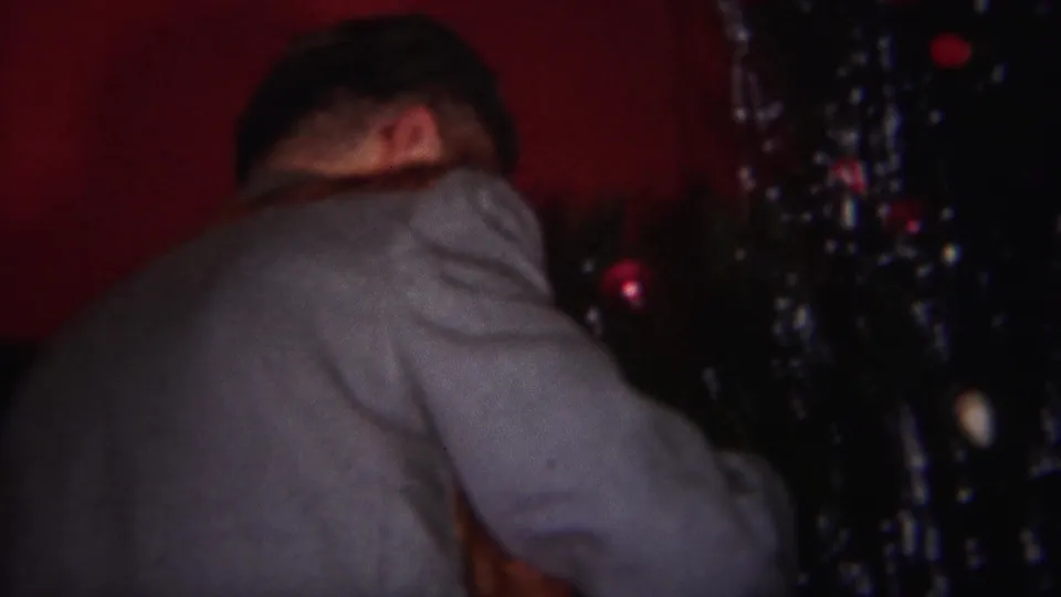 The man turns slightly beside the tinsel-covered Christmas tree, with the red wall and dim home interior visible behind him.