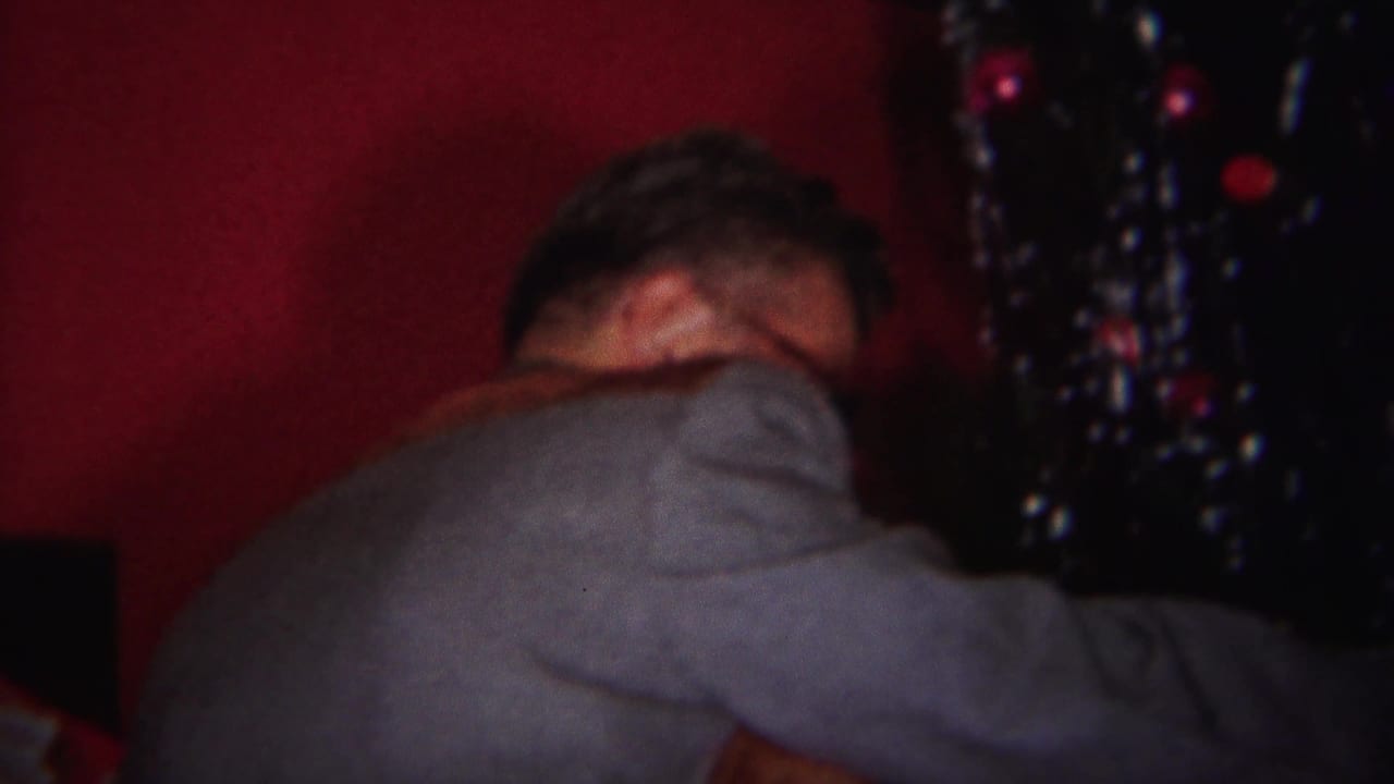 Man bends beside a Christmas tree with ornaments in a dim interior.
