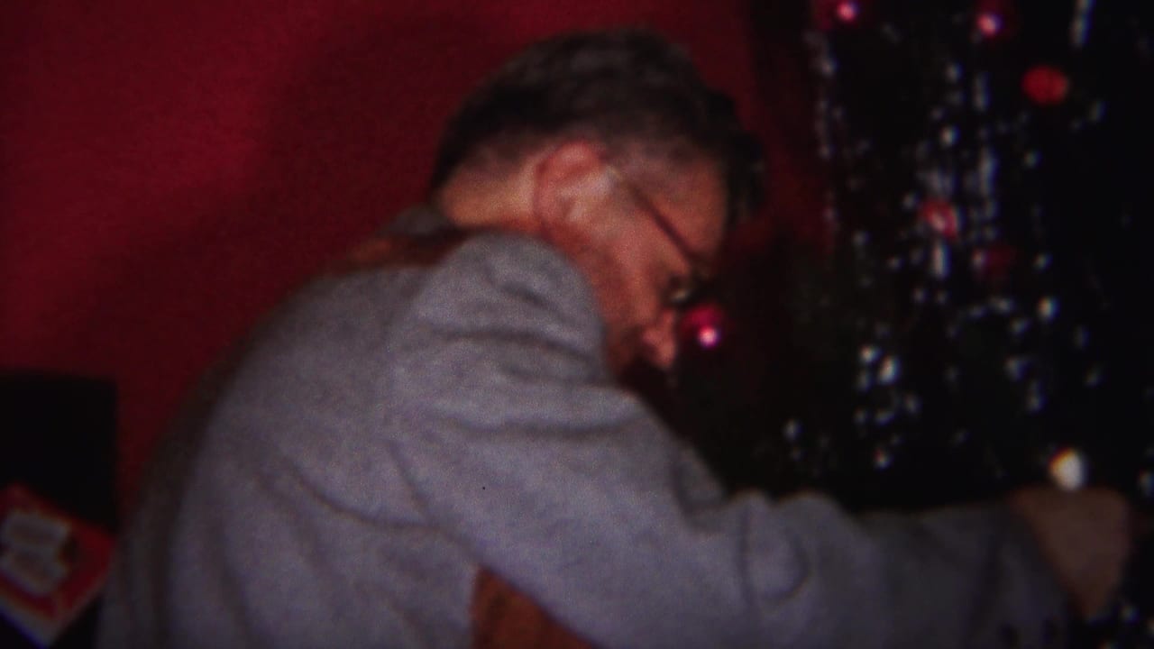 Older man in glasses reaches toward a Christmas tree beside red walls.