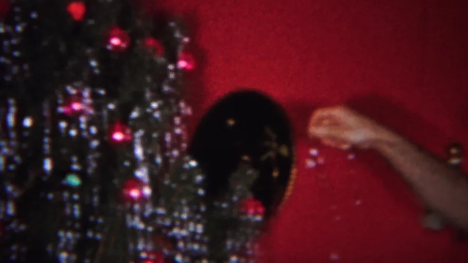A tinsel-covered Christmas tree stands against the red interior wall, with a partial arm entering from the right side of the frame.