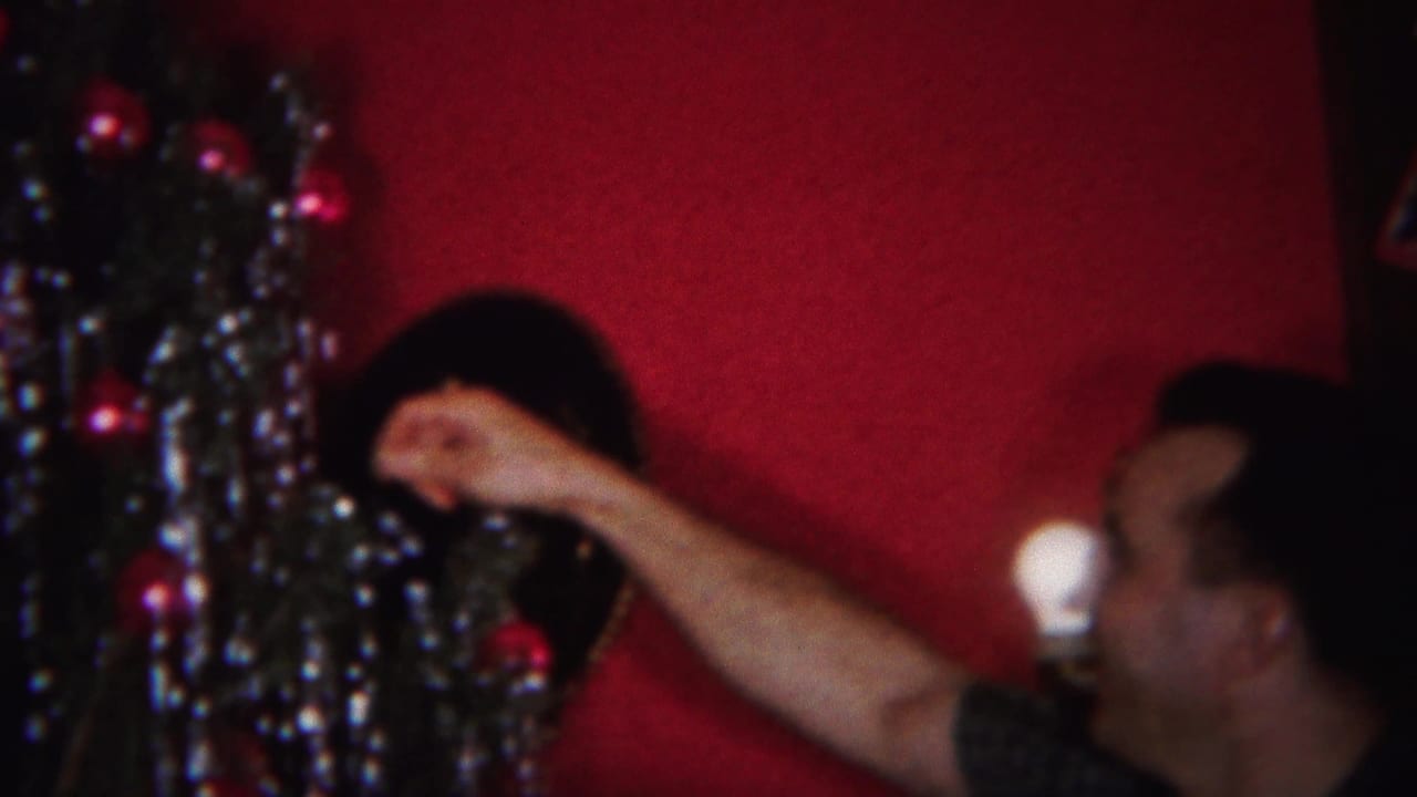 In a 1951 Westville, New Jersey home movie frame, a man reaches toward a Christmas tree with tinsel and ornaments against a red wall.