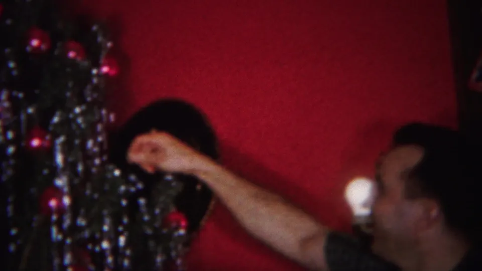 In a 1951 Westville, New Jersey home movie frame, a man reaches toward a Christmas tree with tinsel and ornaments against a red wall.