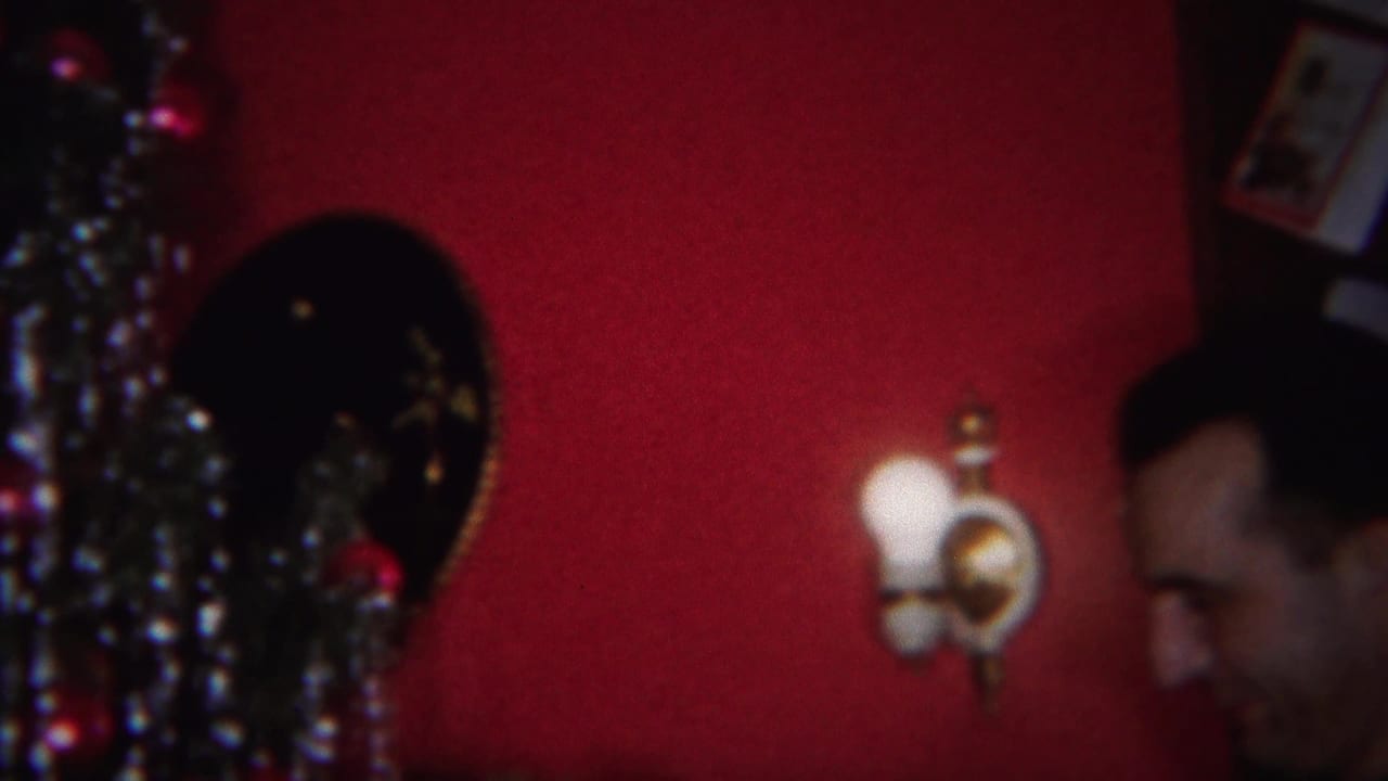 Man near a decorated Christmas tree, with a wall sconce and framed pictures behind him.