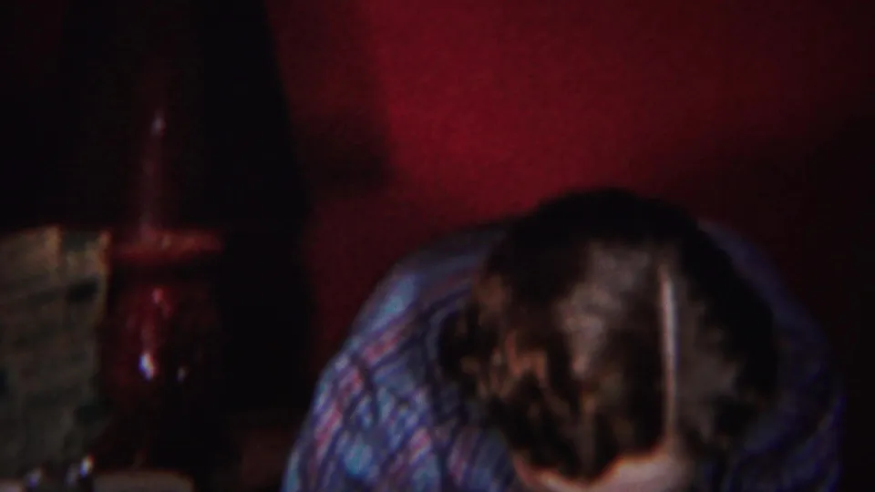 A person in a blue patterned garment bends forward in the dimly lit 1951 Westville interior, with a red wall behind.