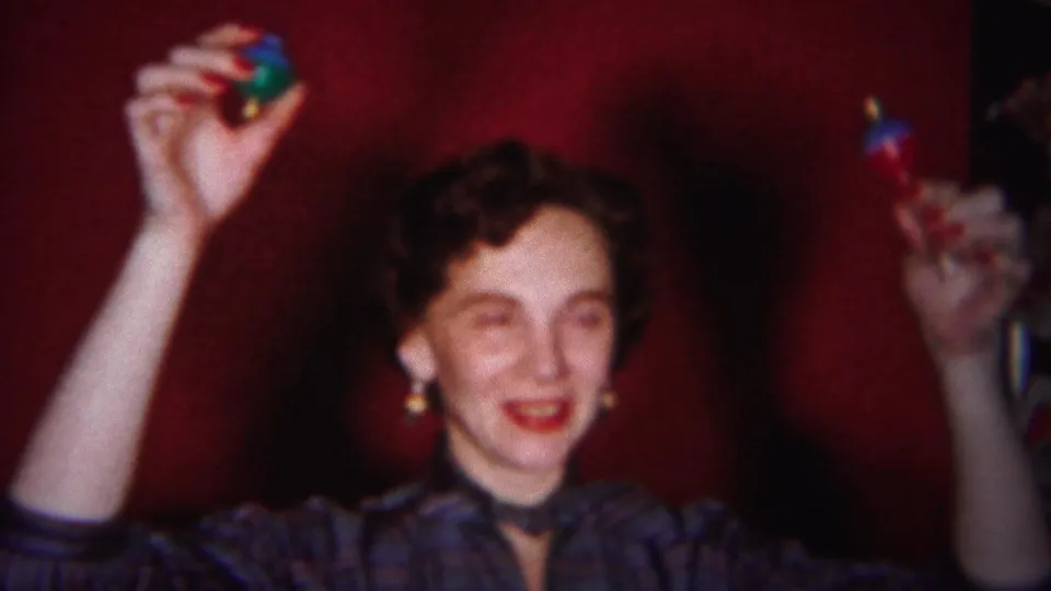 In a 1951 Westville home-movie frame, a woman in a striped collared shirt smiles with both arms raised in a red-walled room.