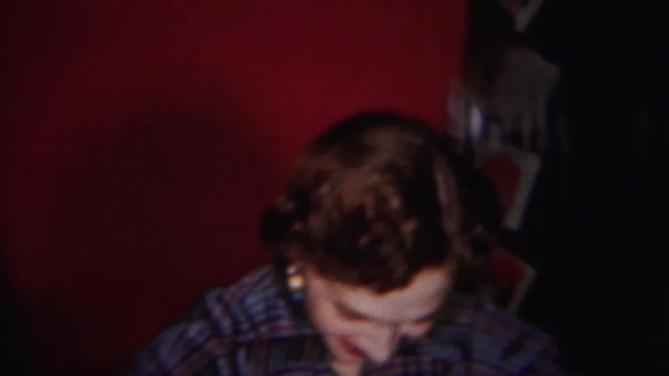 The woman looks downward in the dim red-walled interior, with curled hair and a striped collared shirt visible.