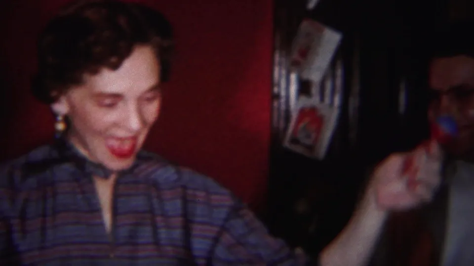 The woman smiles and raises one hand in a small gesture inside the Westville home interior, with framed pictures visible nearby.