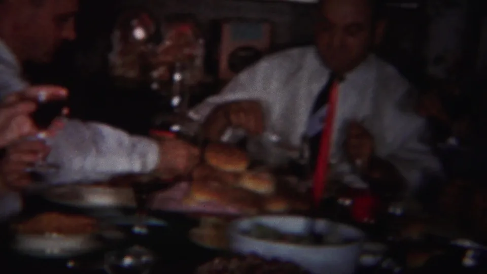 1951 Westville, New Jersey home-movie frame showing adults at a crowded dinner table with bread rolls, plates, and raised glasses.
