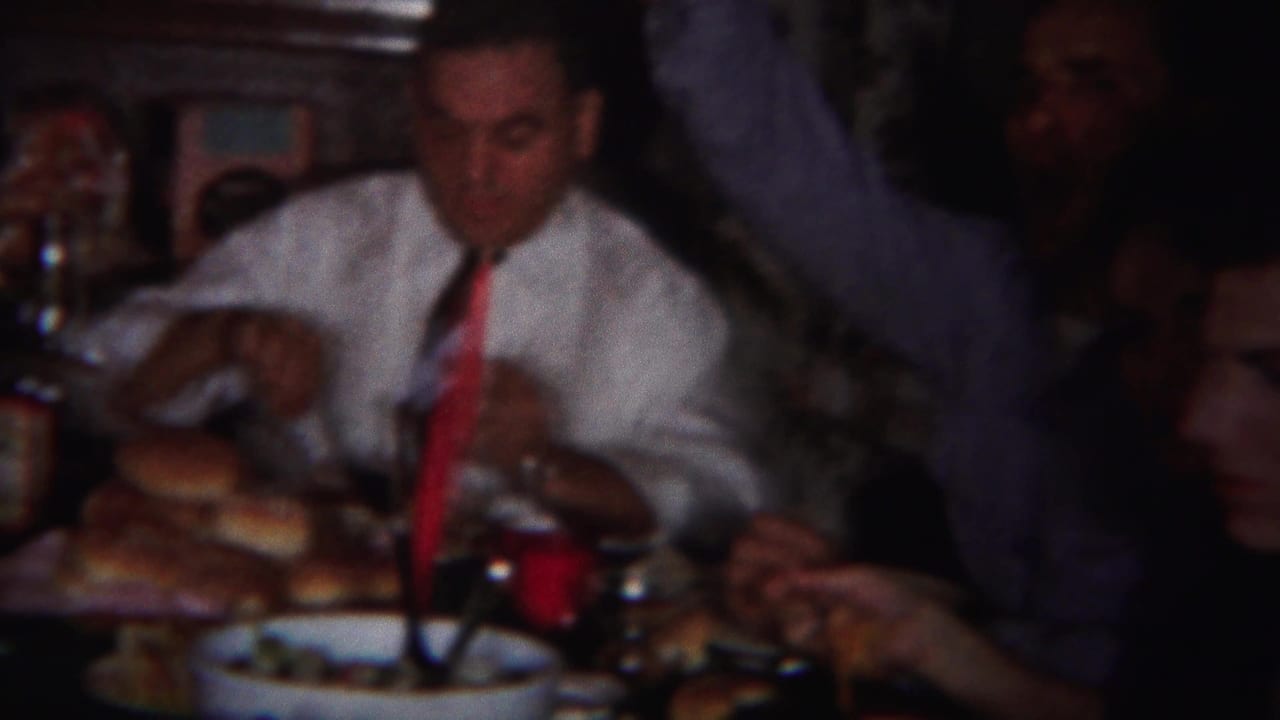 Man in a white shirt and red tie sits beside bread rolls and tableware.