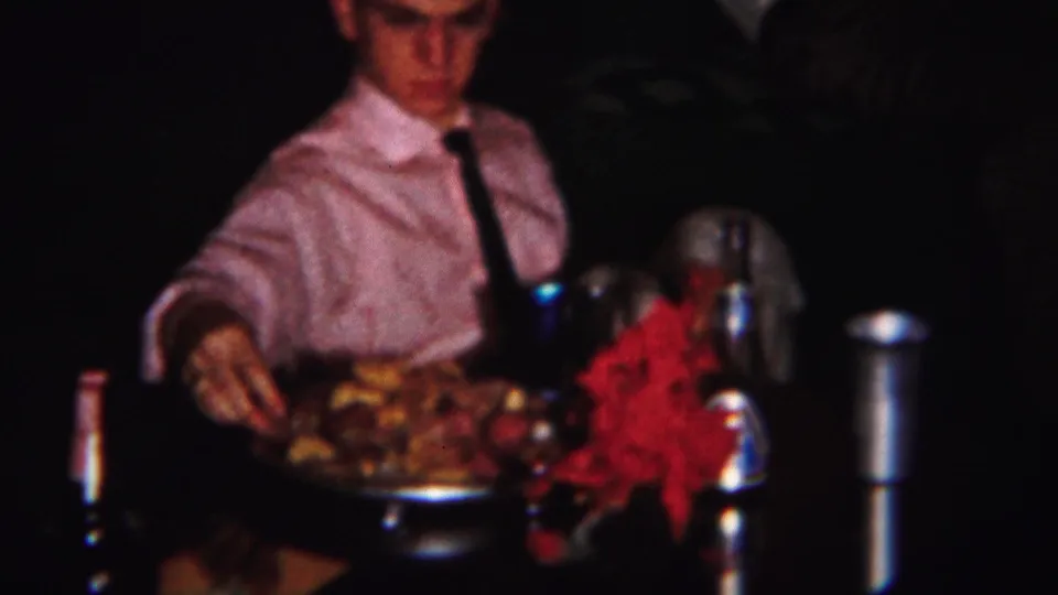 In 1951 Westville home-movie footage, a boy in a pink shirt and tie serves food at a crowded dinner table.