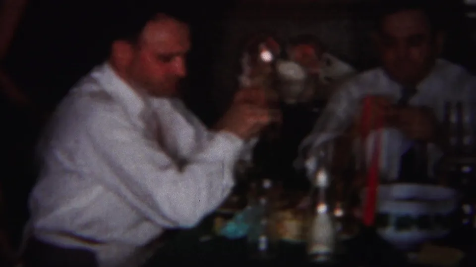 A seated man raises a glass while another sits nearby at the Westville family dinner table in 1951 footage.