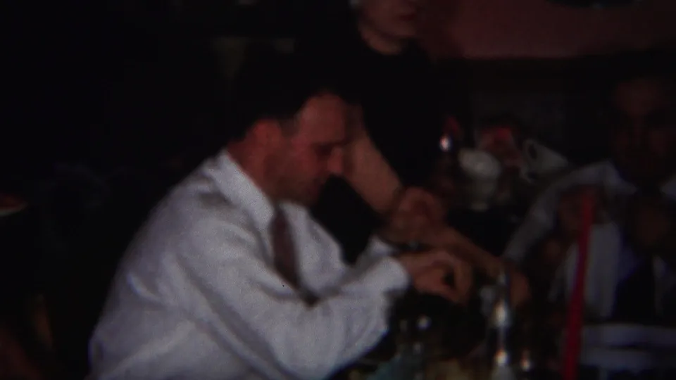 A man in a white shirt leans over the dim Westville dinner table as family members continue eating in 1951 footage.