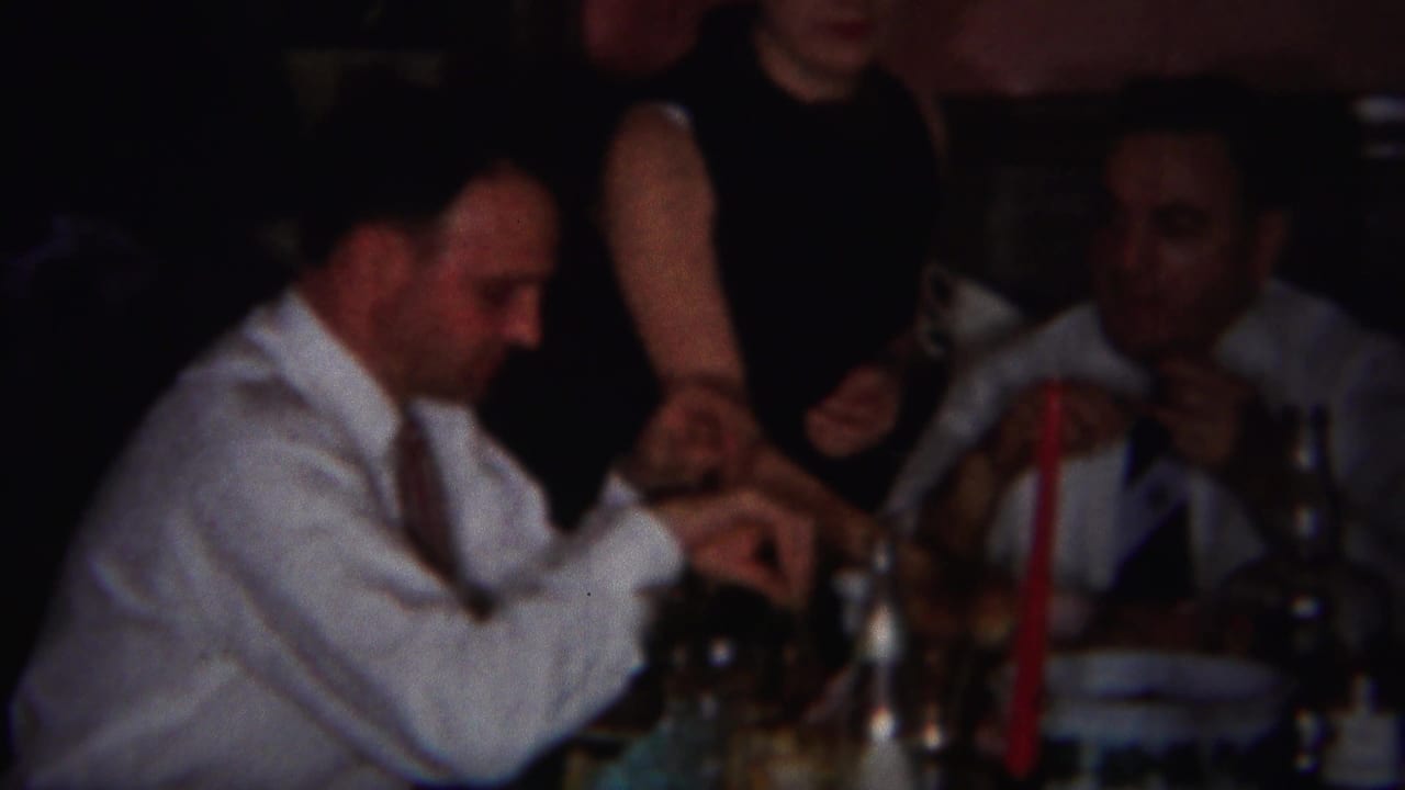 Two men in dress shirts and ties sit at a dining table as a woman stands nearby.