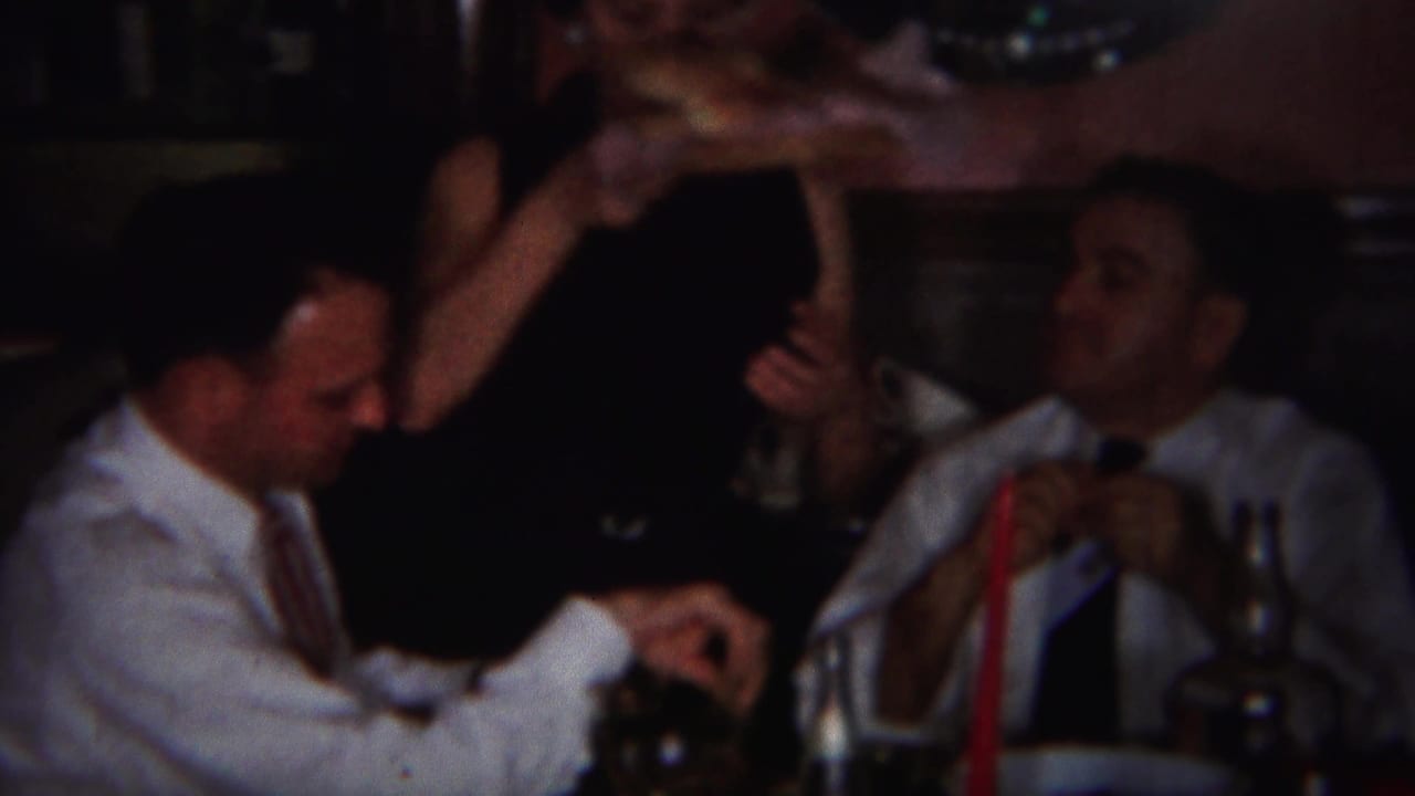 A man in a white shirt reaches toward the table while another man drinks nearby.