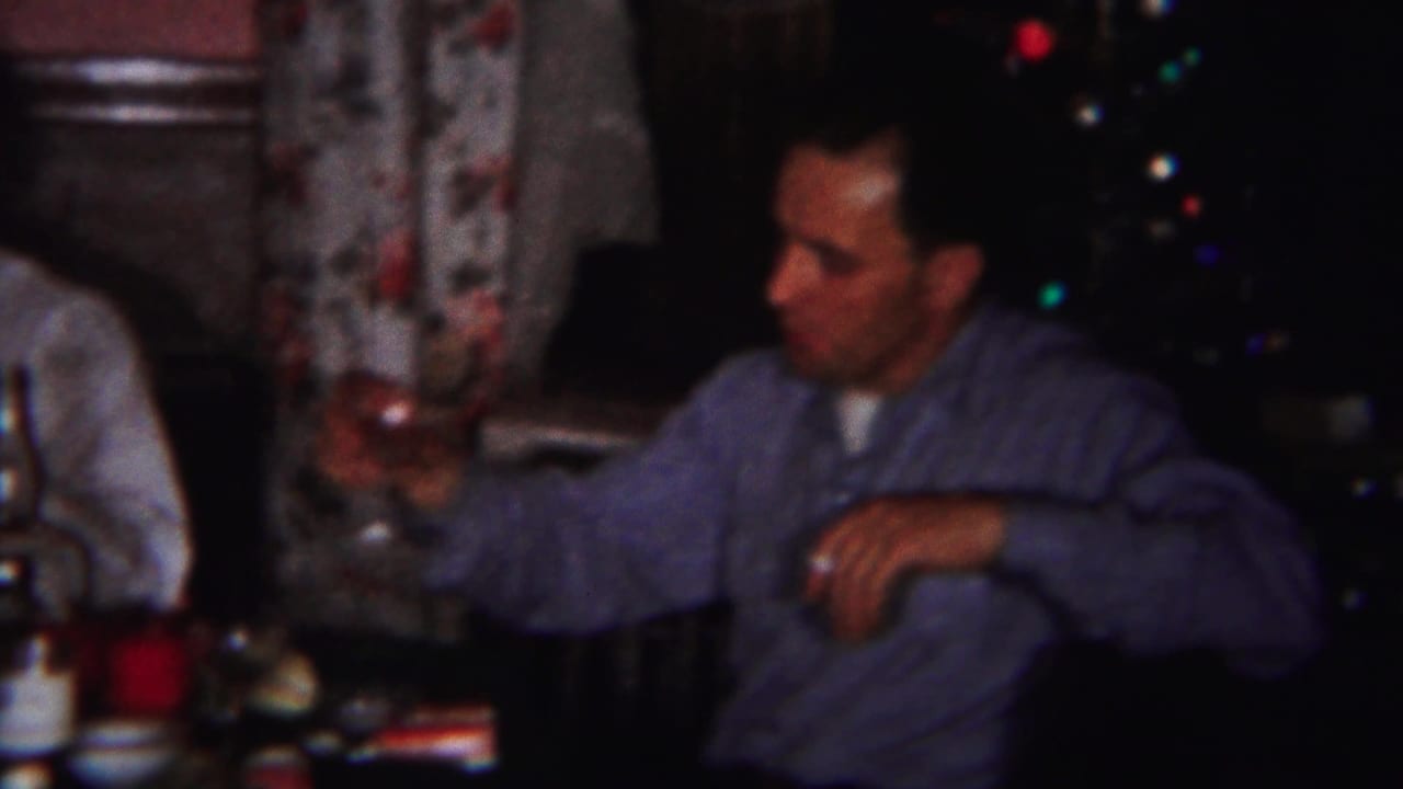 Adult man in a blue shirt holds a drink at a dinner table near a Christmas tree.
