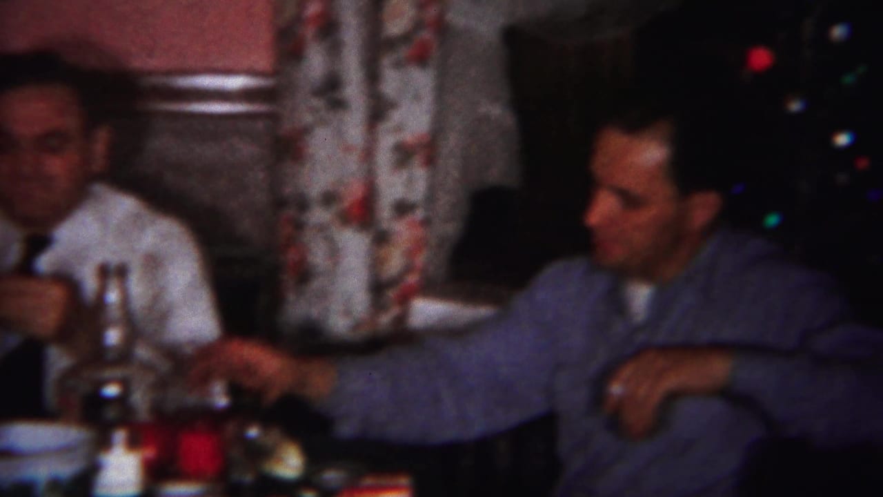Two adult men sit at a table with bottles and glasses near floral curtains and a Christmas tree.