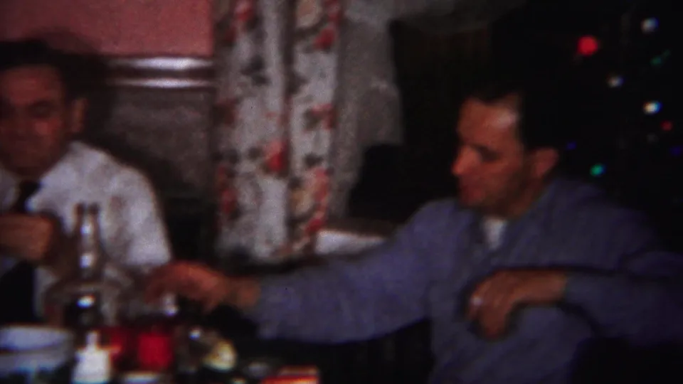 Two adult men sit at a dinner table in a private Westville home, with bottles, glasses, floral curtains, and Christmas tree lights visible.
