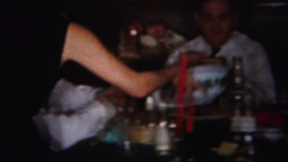 A partially visible woman reaches across a crowded dinner table as an adult man sits behind bottles, glasses, and red candles in the holiday interior.