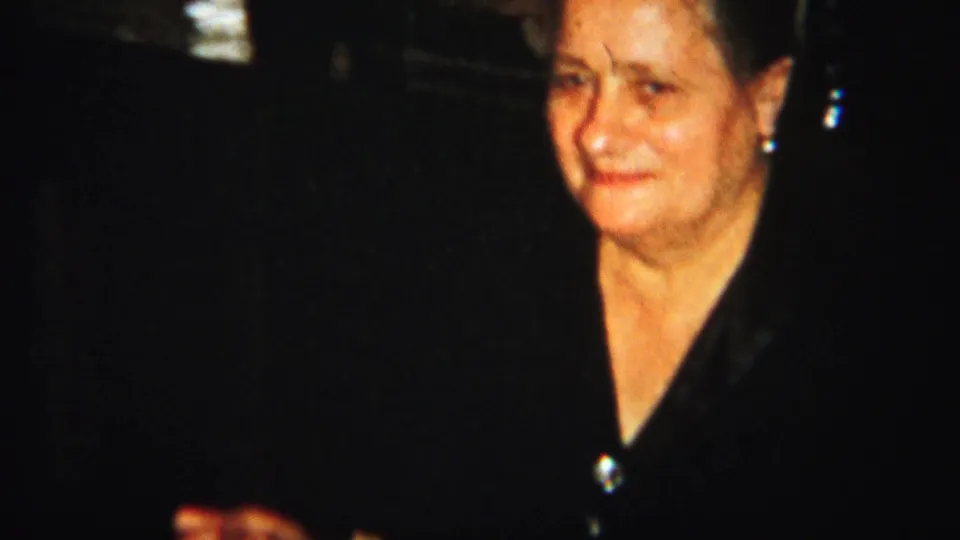 A woman in a dark blouse faces the camera during a 1951 Westville dinner scene, with the room behind her mostly dark.