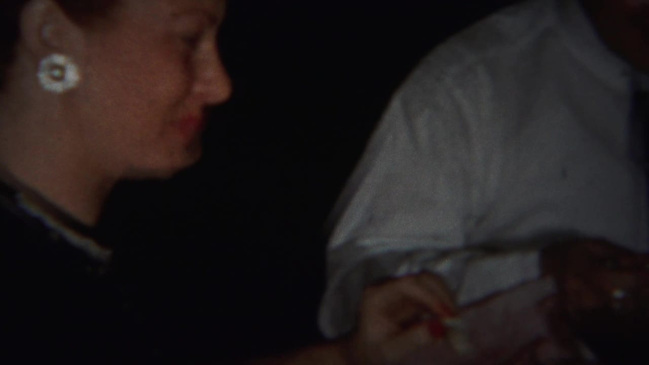 A woman in profile, wearing white flower earrings, appears beside another seated adult in this 1951 Westville home-movie frame.