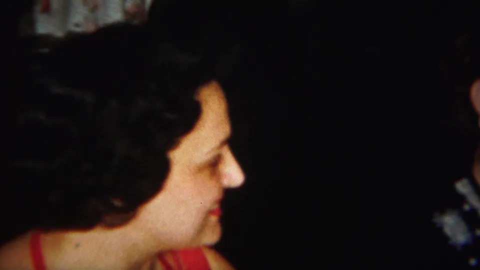 The woman remains turned right and looks slightly downward, with soft focus and grain from the 8mm home movie.