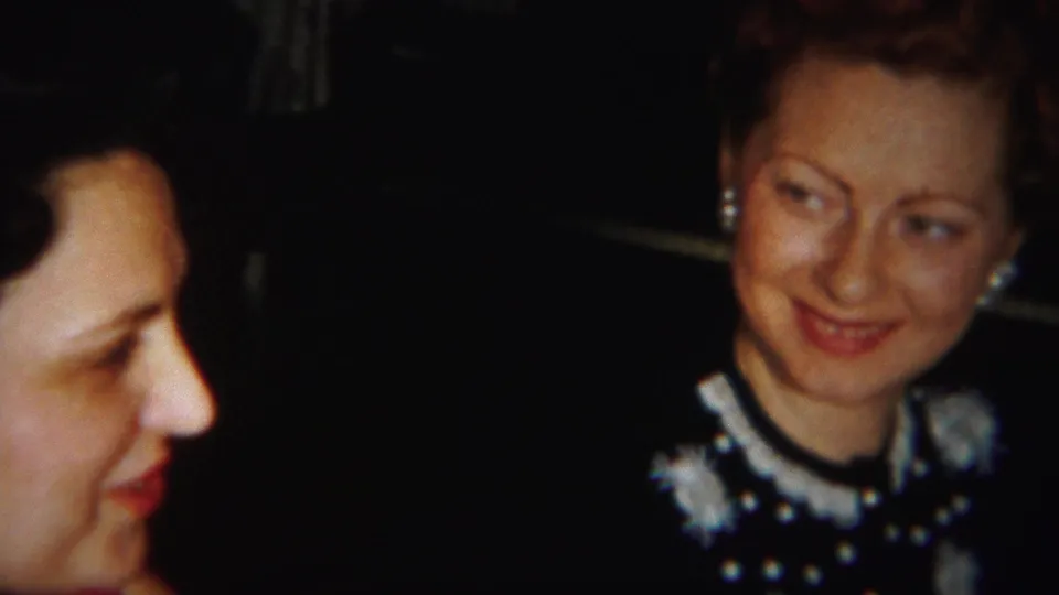 Two adult women are framed closely indoors, with one partly cropped at left and another in a dark dress with white collar details smiling sideways.