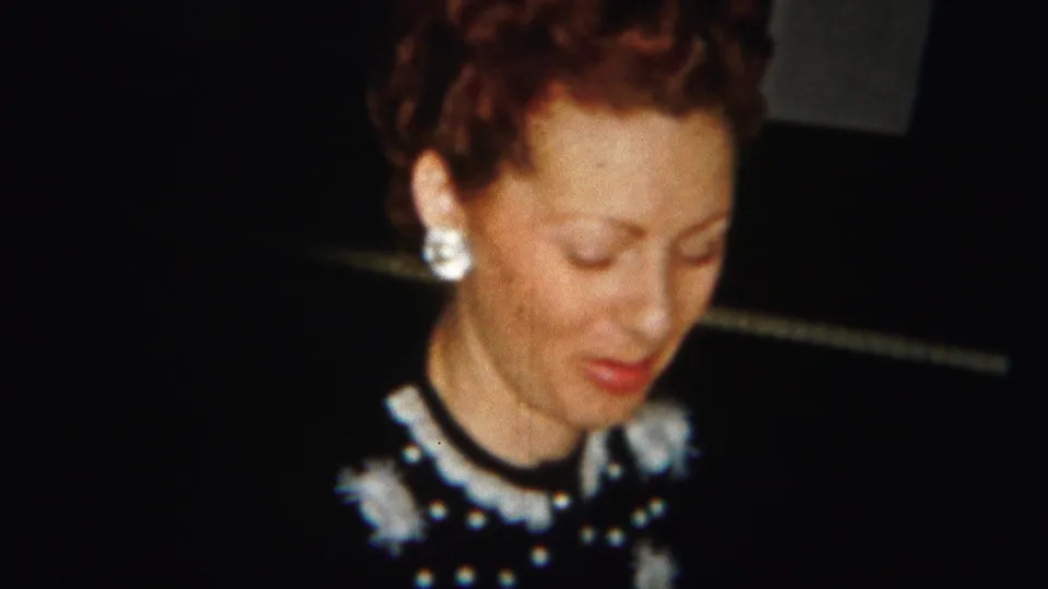 A woman in a dark dress with white collar-like embellishment and earrings looks down in close 1951 Westville home-movie footage.