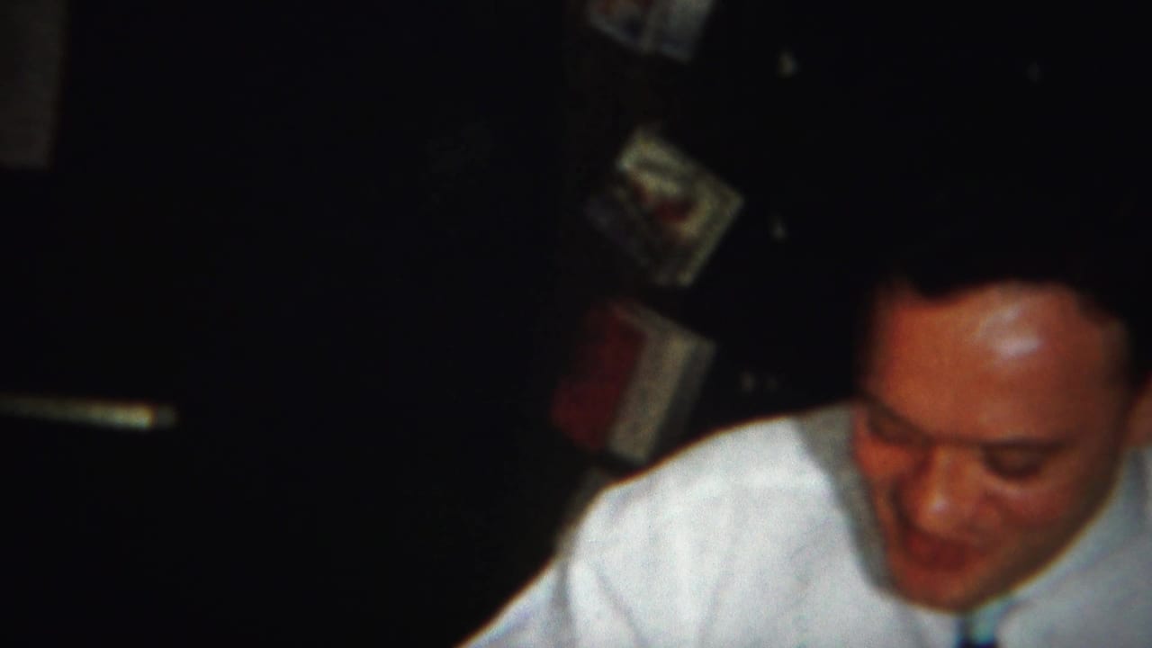 A man in a white shirt and dark tie looks down during the Westville, New Jersey home-movie dinner scene.