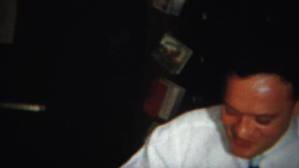 A man in a white shirt and dark tie looks down during the Westville, New Jersey home-movie dinner scene.