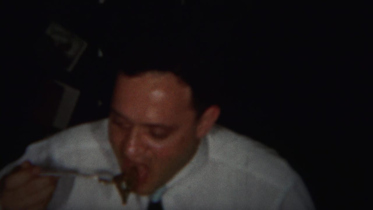 Man in a white shirt and tie takes a bite from a fork in a dim room.