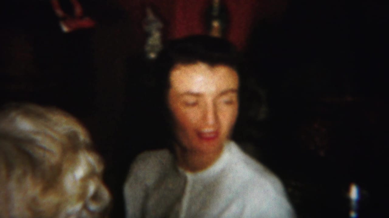 Woman in a light sweater seated indoors beside another person in dim light.