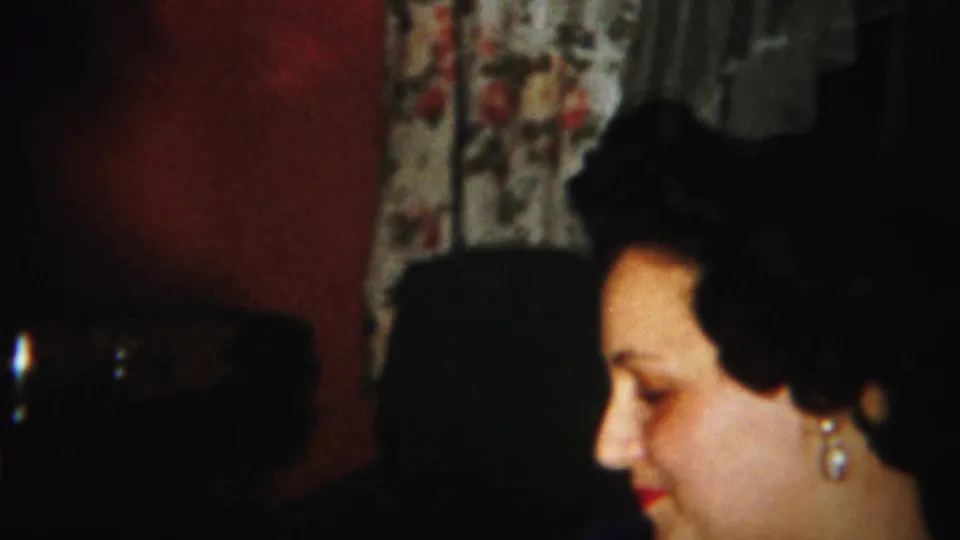 A woman in profile sits near floral curtains in a dim 1951 Westville home-movie interior.