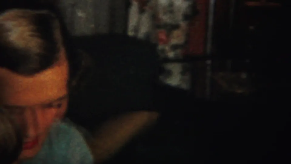 Partly obscured adult face at the left edge of a dim 1951 Westville indoor home-movie frame, with a foreground head and dark furnishings.