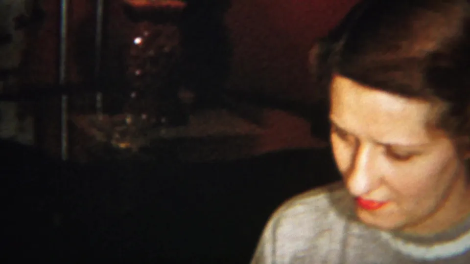 Adult woman with red lipstick, pale top, and neatly styled hair looks downward in a dim Westville, New Jersey home interior.