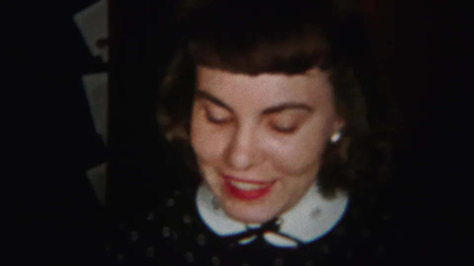 A close home-movie frame shows the woman looking down with a smile, her earrings, white collar, bow, and dark polka-dot dress visible.
