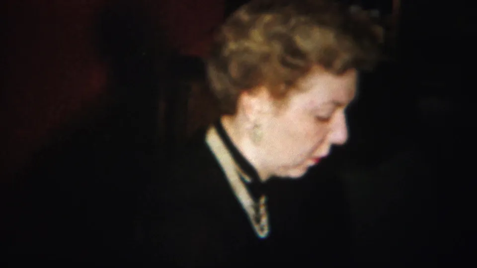 An older woman with curled light hair appears in profile, looking downward in the same dim indoor 1951 Westville home-movie setting.
