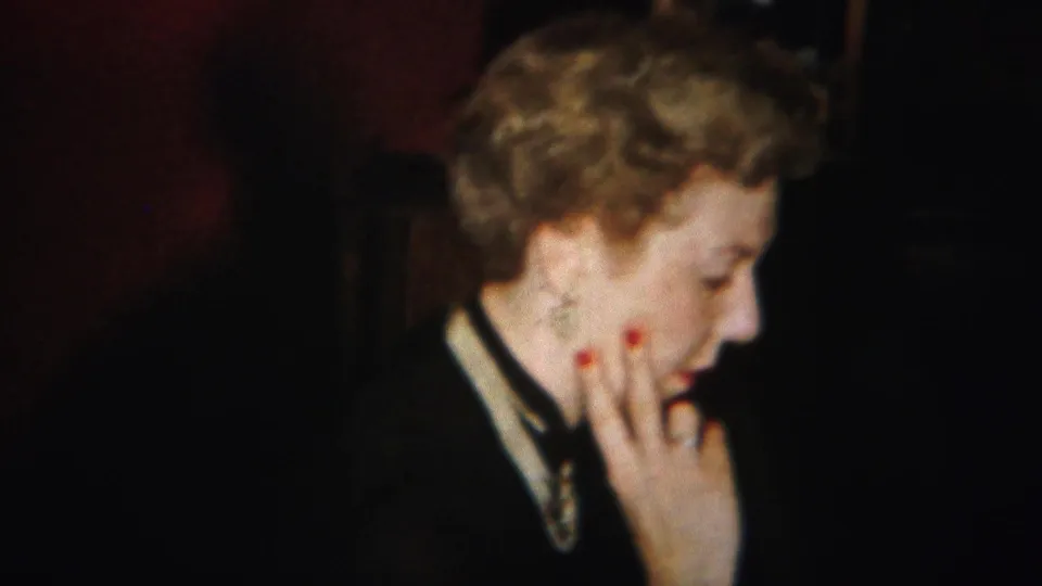 In the 1951 home-movie interior, the woman raises a hand with red nail polish to her cheek while looking down.