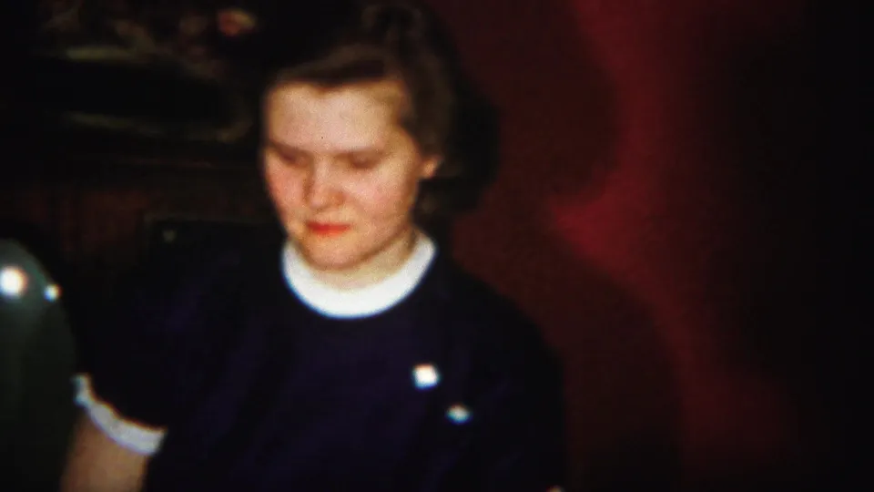 The home-movie frame shows the seated young woman near dark furniture and red walls during the indoor gathering.